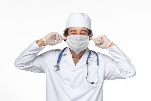 外套正面图：男医生穿着医疗服 戴着口罩以防感染冠状病毒——在白墙冠状病毒大流行的药物上闭上耳朵视野防护套装