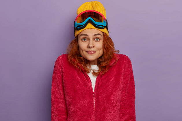 欢呼快乐的年轻红发女士穿着暖和的衣服 头戴滑雪镜 在紫色背景下摆姿势滑雪板酒窝冬天