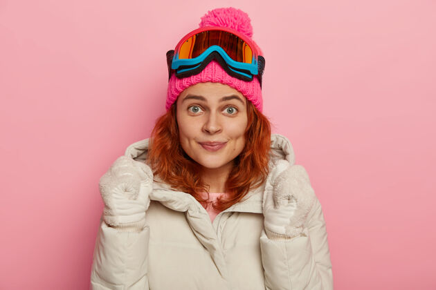 眼镜快乐的滑板运动员举手欢呼 穿着冬衣 戴着手套 戴着滑雪面具 愉快地看着镜头 对着玫瑰色的工作室墙壁摆姿势复制空间喜悦室内