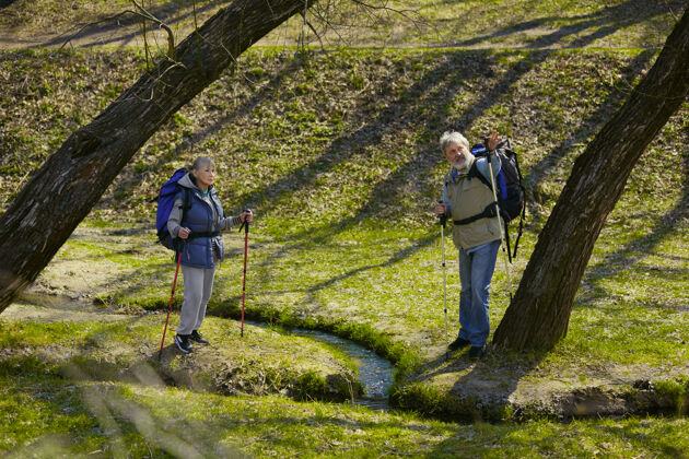 聚在一起幸福的回忆阳光明媚的日子里 一对穿着旅游服装的老年夫妇在小溪边的绿色草坪上散步老年享受老年人