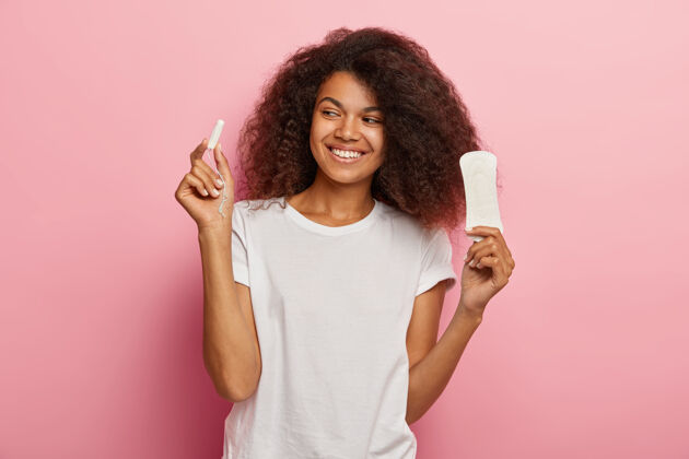 高兴快乐的非洲裔美国妇女拿着卫生棉条和卫生巾 穿着白色t恤 隔离在粉红色的墙上女人 经前综合症室内美式月经