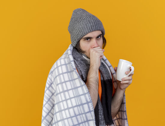 生病咳嗽的年轻病人戴着冬天的帽子 围巾裹着格子布 手里拿着一杯橘黄色的茶冬天男人年轻人