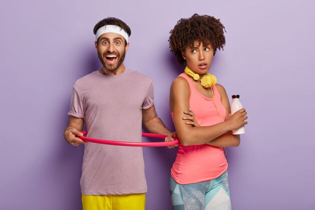 女人室内拍摄笑脸男子旋转呼啦圈 身着紫色t恤 身材匀称 非洲裔女子向后站着 拿着一瓶淡水 隔着紫色的墙壁健康的生活方式健康身体紫罗兰