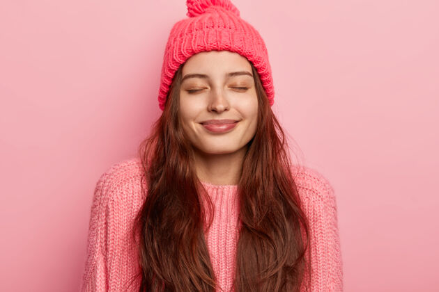 罗西快乐的长发快乐的白种女人的照片闭上眼睛 微笑温柔 有健康的皮肤 戴着温暖的针织帽子和套头衫 在粉色背景下摆姿势 想象一些美好的事情服装女人年轻
