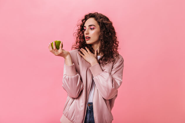 深色头发穿着粉红色夹克的女人在孤立的背景下看着苹果积极时尚健康