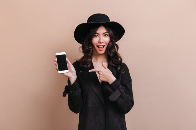摄影棚拍摄亚洲女人用手指着智能手机 屏幕是空白的一个情绪激动的穿着黑色外套的韩国女人的摄影棚镜头快乐帽子时尚