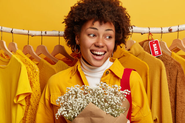 消费者黑色星期五和降价概念积极的卷发女人欢喜商店五折优惠 可以买很多衣服不多的钱 站在展示台附近与黄色的衣服 携带鲜花牙齿高兴微笑