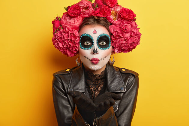油漆为墨西哥嘉年华做准备的女性骷髅肖像 吹着飞吻 化着头骨妆 穿着黑色夹克 看起来很吓人 隔离在黄色背景下人体彩绘和面部艺术身体服装棕榈