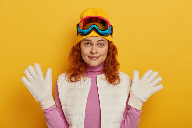 运动员开朗自然的女人戴着白手套举手 戴着滑雪板眼镜 享受阳光明媚的冬日 开心地看着镜头 在黄色背景下摆姿势拿着女运动员红发