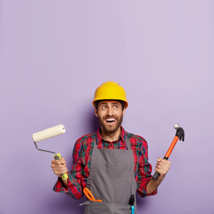 头盔图为兴高采烈的男建筑工人或装饰工人手持铁锤和油漆辊 准备粉刷墙壁和修补 表情愉快 抬头看去建筑工黄色锤子