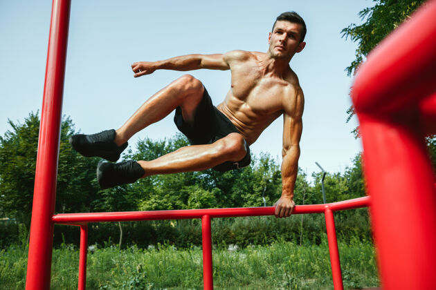 肌肉在阳光明媚的夏日 一个年轻肌肉发达 赤膊上阵的白人男子在操场上跳过单杠锻炼水平区域