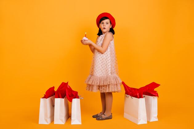 女孩带着购物袋的小女孩的完整镜头戴着红色法国贝雷帽的可爱的孩子被隔离在黄色的墙上商店购物者未成年