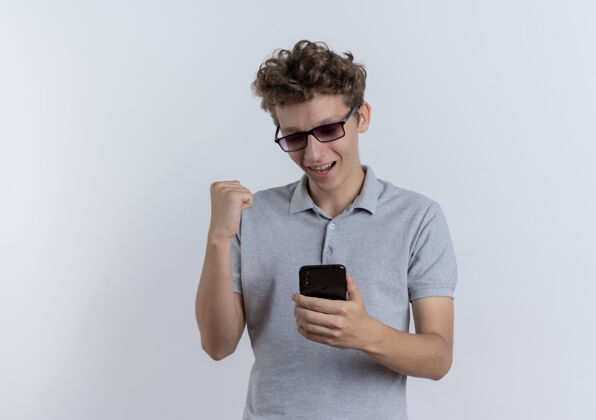 年轻戴着黑眼镜 身穿灰色马球衫的年轻人看着智能手机屏幕 握紧拳头 站在白色的墙上 快乐而兴奋兴奋马球手机
