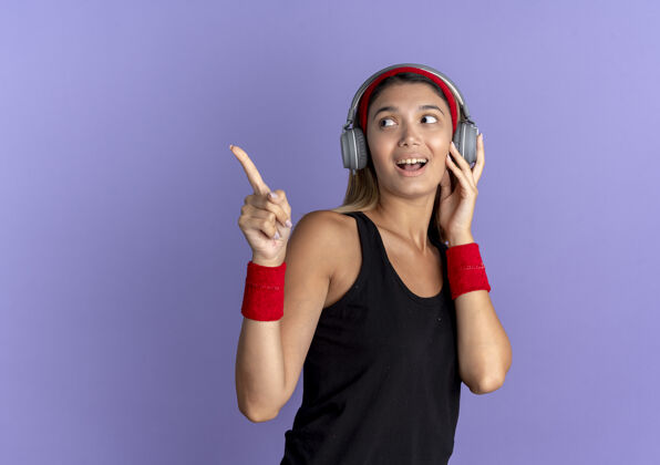 女孩身穿黑色运动服 头戴红色耳机的年轻健身女孩微笑着用手指着站在蓝色墙上的一边耳机运动装头带