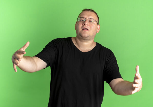 男人戴眼镜的胖男人穿着黑色t恤做欢迎手势张开双手站在绿色的墙上手势宽手
