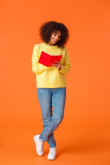 日程安排全长垂直拍摄梦幻可爱的非洲裔美国女学生 留着非洲发型 穿着休闲服 上大学 做笔记 站在橙色的墙上 微笑着在笔记本上写字全长笔记本写作