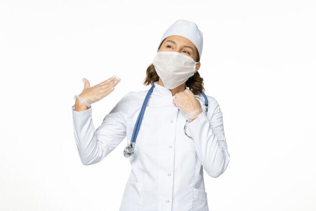 专业正面图年轻女医生由于白色表面有冠状病毒 戴着无菌口罩和手套听诊器女性医生