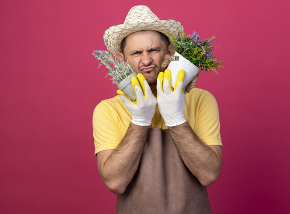 不高兴年轻的园丁戴着工作手套 穿着连体衣 戴着帽子 手里拿着盆栽植物 感到困惑和不快粉色锅站着