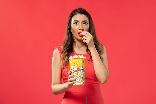 爆米花正面图穿红衬衫的年轻女性在红桌子上吃爆米花看电影电影前面吃