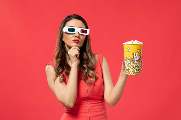 性感正面图戴着d型太阳镜拿着爆米花的年轻女性在浅红色的表面思考前面电影举行