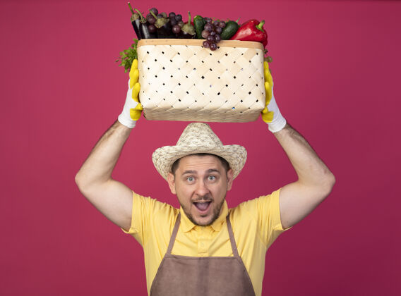 帽子年轻的园丁戴着工作手套 穿着连体衣 戴着帽子 头上抱着装满蔬菜的箱子 高兴而兴奋地笑着粉色手套满的