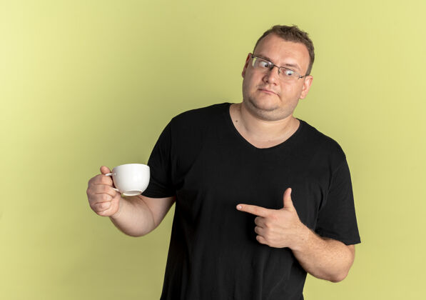 咖啡戴眼镜的超重男子身穿黑色t恤 手拿咖啡杯 手指指着咖啡杯 困惑地站在浅色的墙上望向一边穿超重眼镜