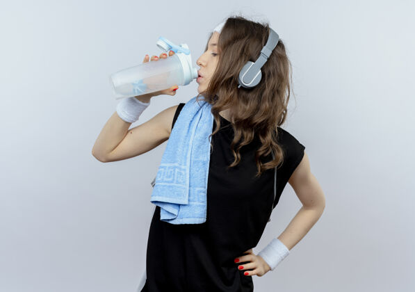 毛巾穿着黑色运动服的年轻健身女孩 戴着头带和耳机 肩上放着毛巾 锻炼后站在白墙上喝水运动服健身耳机