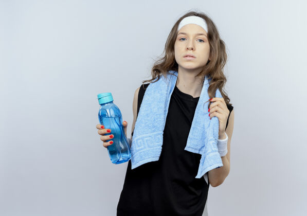 瓶子穿着黑色运动服的年轻健身女孩 头上戴着头巾 脖子上围着毛巾 手里拿着一瓶水 严肃的脸站在白色的墙上看脖子脸