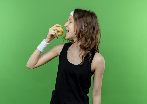 女孩身穿黑色运动服 头箍咬着绿苹果站在绿墙上的年轻健身女孩运动装健身年轻
