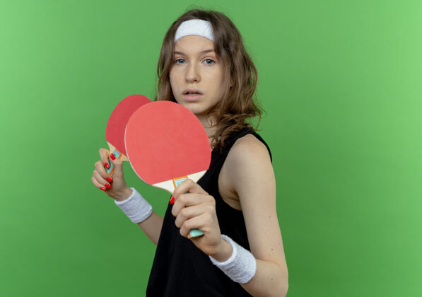 年轻身穿黑色运动服 头戴钢带 手持两个乒乓球拍的年轻健身女孩 严肃的脸站在绿色的墙上女孩抱站