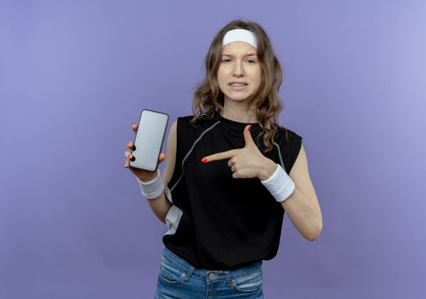 手指身穿黑色运动服 头戴智能手机 食指指着智能手机的年轻健身女孩站在蓝色的墙上 看起来很自信立场移动显示