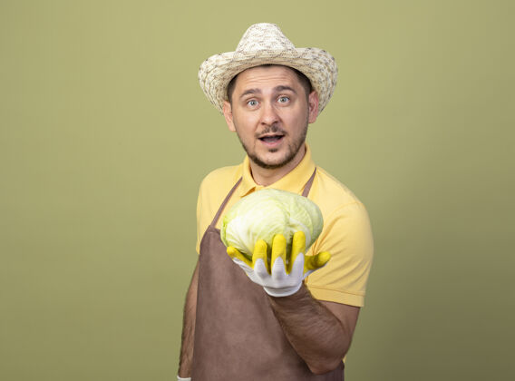 卷心菜年轻的园丁戴着工作手套 穿着连体衣 戴着帽子 在镜头前露出洋白菜的笑脸站着微笑花园