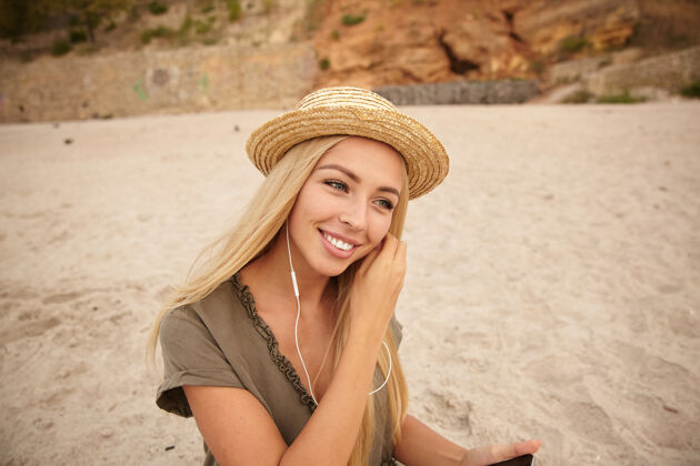 表情积极的年轻漂亮的白头女子戴着背带帽 一边看着前方 一边高兴地微笑着 一边用耳机听音乐 被隔离在海边头发海滩帽子