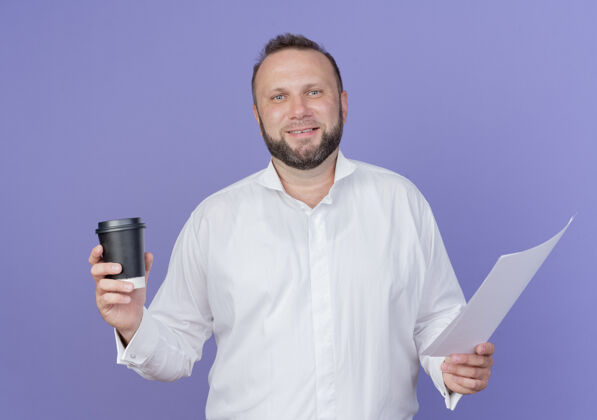 男人一个留着胡子的男人 穿着白衬衫 手里拿着咖啡杯 站在蓝色的墙上 面带微笑微笑拿着胡子