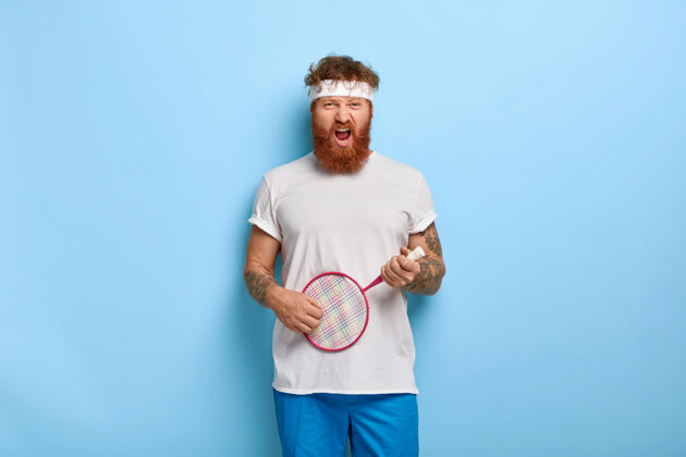 人有趣的红头发网球运动员拿着球拍对着蓝色的墙摆姿势男人网球游戏