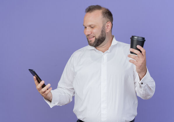 男人一个留着胡子的男人穿着白衬衫 手里拿着咖啡杯 站在蓝色的墙上微笑着看着他的智能手机屏幕手机咖啡拿着