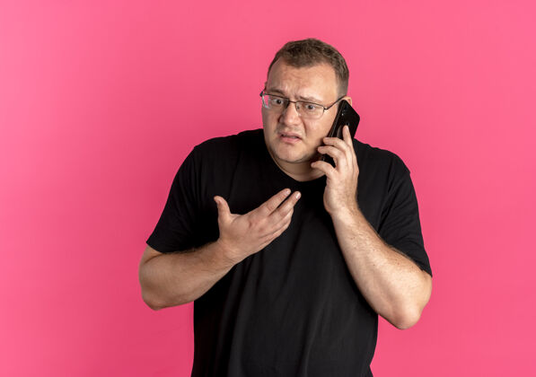 穿一个戴眼镜 身穿黑色t恤的超重男子站在粉红色的墙上 一边打手机一边显得很困惑电话困惑眼镜