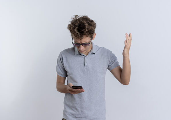 看一个穿着灰色马球衫的年轻人站在白色的墙上 看着他的智能手机屏幕举起的手 感到困惑手年轻举起