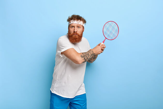 红发坚定的红发网球运动员拿着球拍对着蓝色的墙壁摆姿势人运动活跃