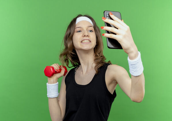 年轻身穿黑色运动服的年轻健身女孩戴着头巾 手里拿着哑铃 站在绿色的墙上用智能手机自拍健身举行手
