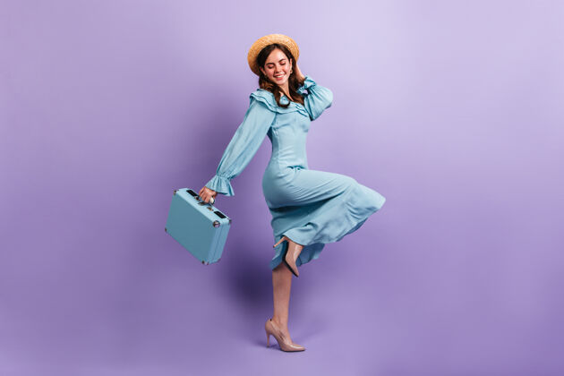 蓝色连衣裙穿着丝绸迷笛裙的可爱的旅行者在紫色的墙上愉快地摆着姿势一个戴草帽带着手提箱的女孩的全长镜头优雅女孩黑发
