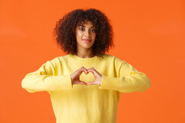 肖像体贴温柔 可爱浪漫的非裔美籍女性表达自己的情感 展示心形 在情人节表白爱意 站在橘色的墙上开心微笑女孩举行