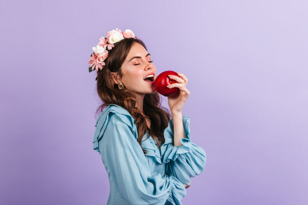 蓝色连衣裙戴着花冠的迷人女士咬着多汁的苹果紫丁香墙上有一个穿蓝色上衣的女孩的画像脸成人肖像