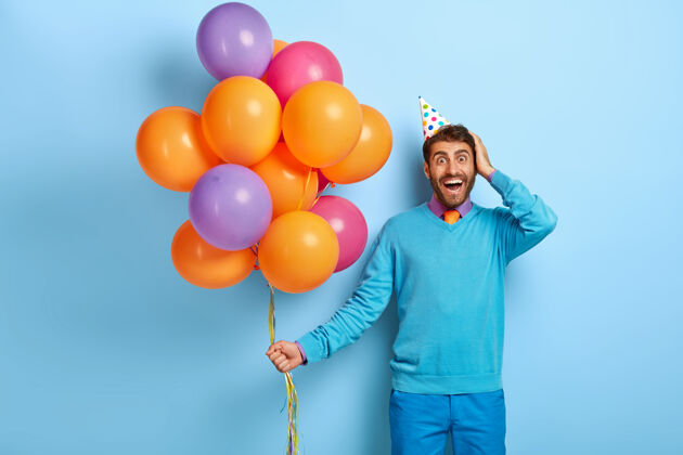 帽子摄影棚拍摄了一个兴奋的家伙戴着生日帽和气球 穿着蓝色毛衣摆姿势姿势满意气球