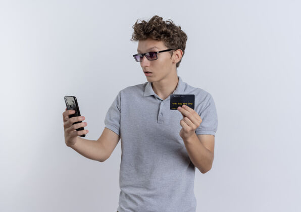 信用一个穿着灰色马球衫的年轻人看着他的智能手机屏幕 屏幕上显示信用卡正站在白色的墙壁上困惑困惑年轻屏幕