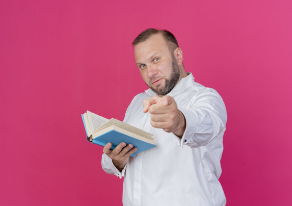 微笑留着胡子的男人穿着白衬衫 拿着打开的书 食指微笑着站在粉红色的墙上手指目录指着
