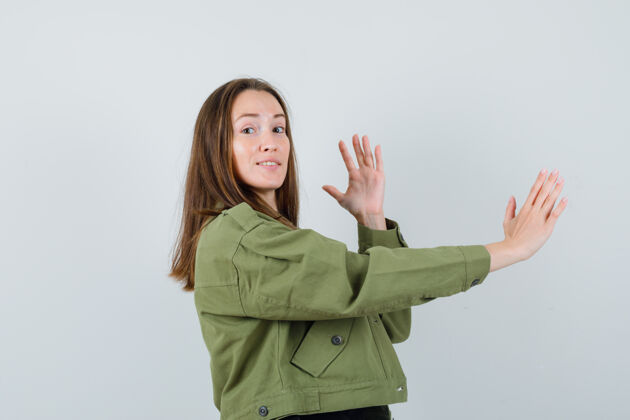肖像穿着绿色夹克的年轻女子举起双手 手掌张开放在一边 小心地看着前方成人女人提高