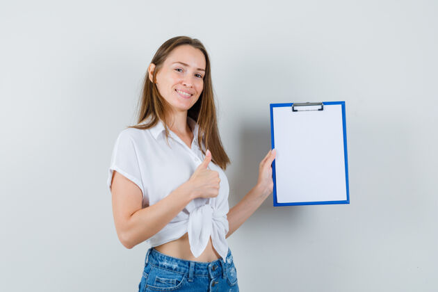 成人穿着白衬衫的年轻女士拿着空白的剪贴板 拇指朝上 正视图穿着空白微笑