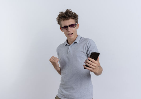 高兴身穿灰色马球衫的年轻人看着智能手机屏幕握紧拳头 为自己站在白墙上的成功而欢欣鼓舞年轻屏幕移动