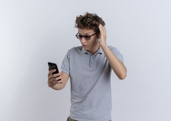 困惑一个穿着灰色马球衫的年轻人站在白色的墙上看着他的智能手机屏幕感到困惑年轻屏幕站